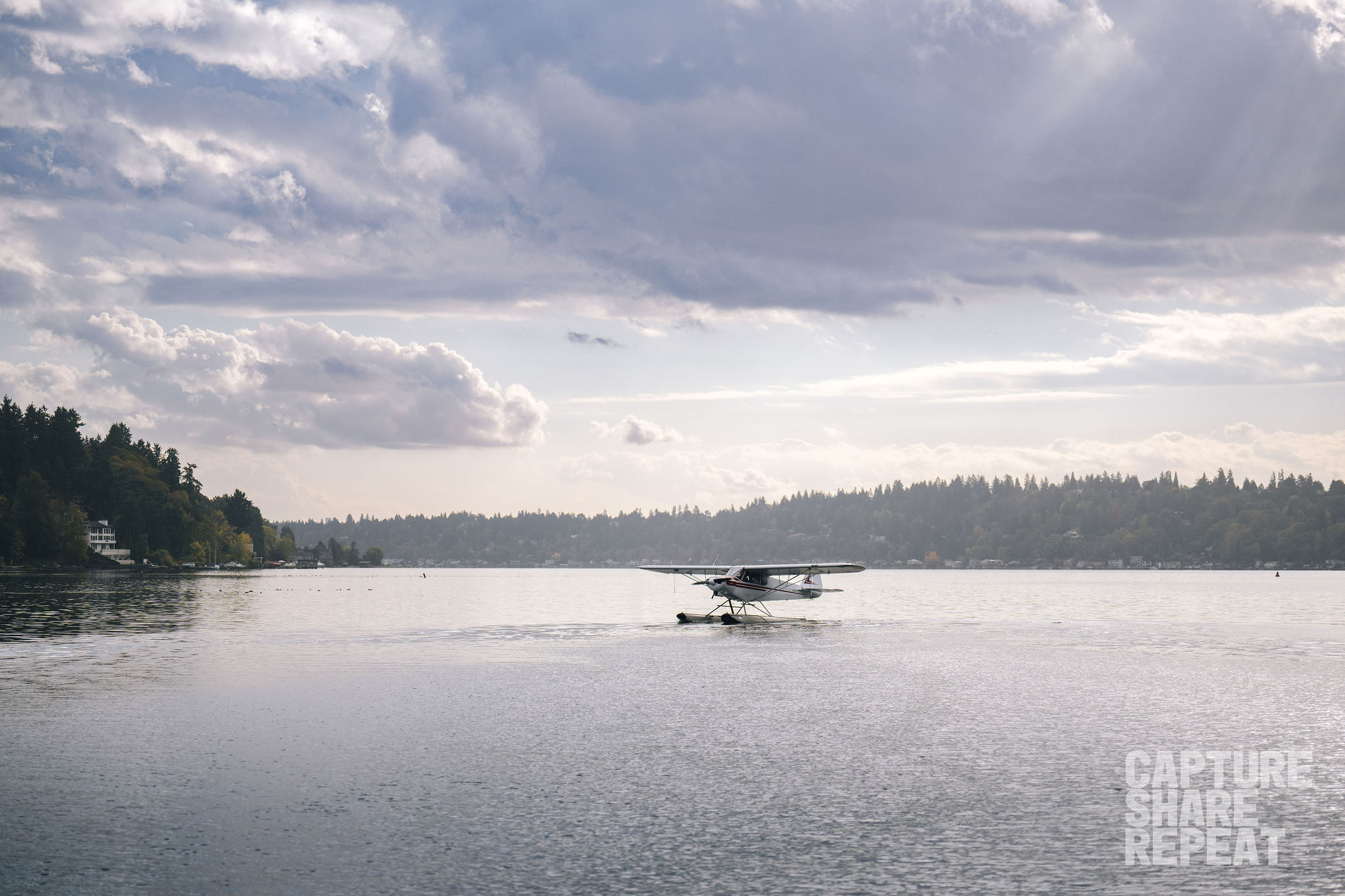 Seaplane landing on lake with rays of sunshine peering through a partly cloudy sky.