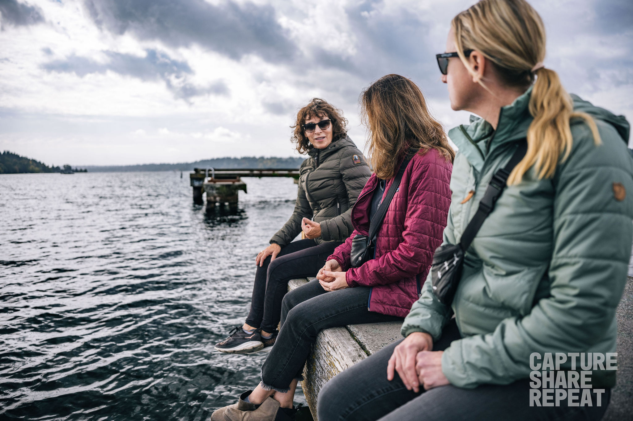 Three women sitting on the edge of a dock chatting with feet dangling over the water.