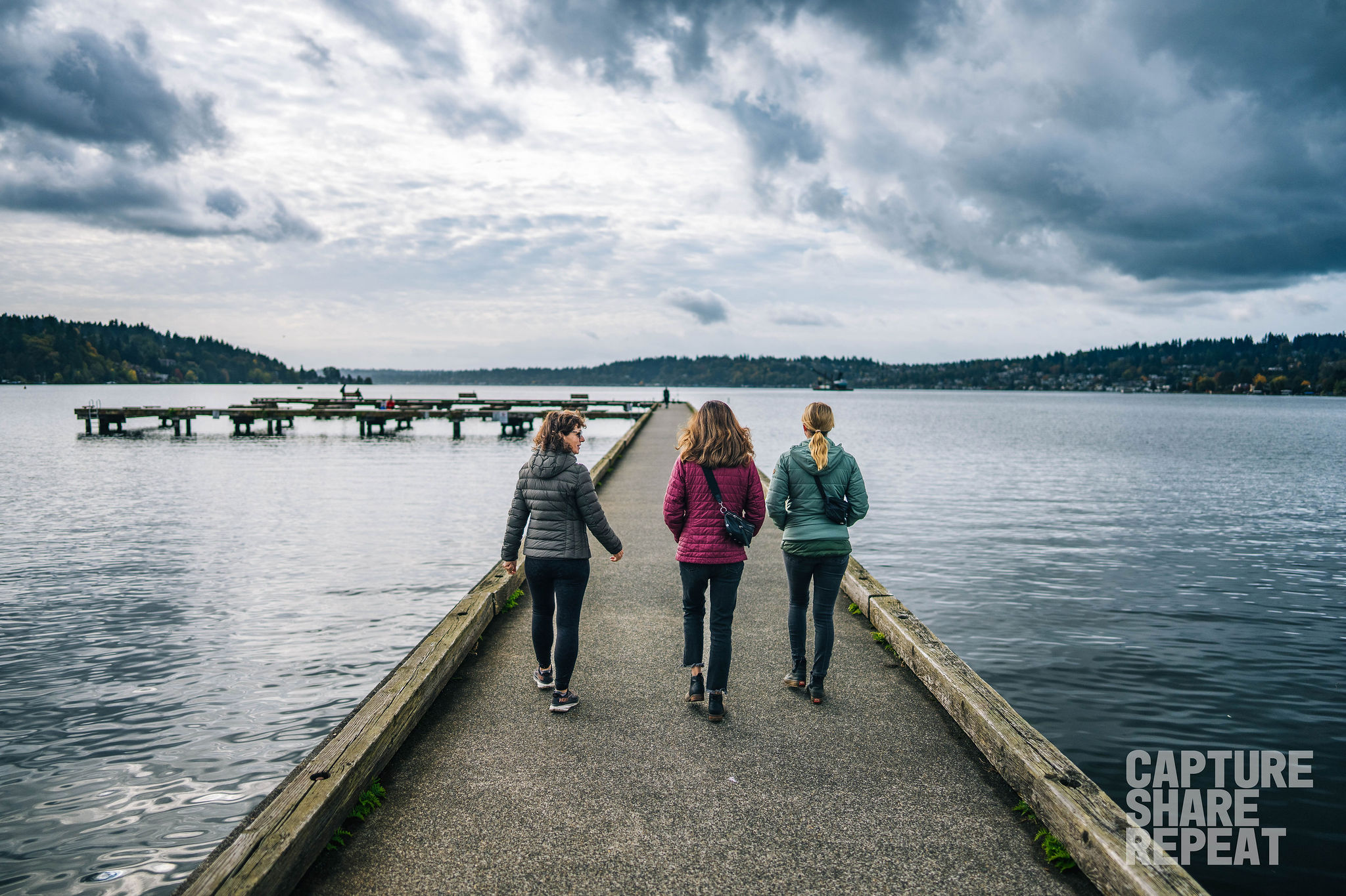 Three women walking out on a dock over a lake with trees on the horizon and cloudy skies.