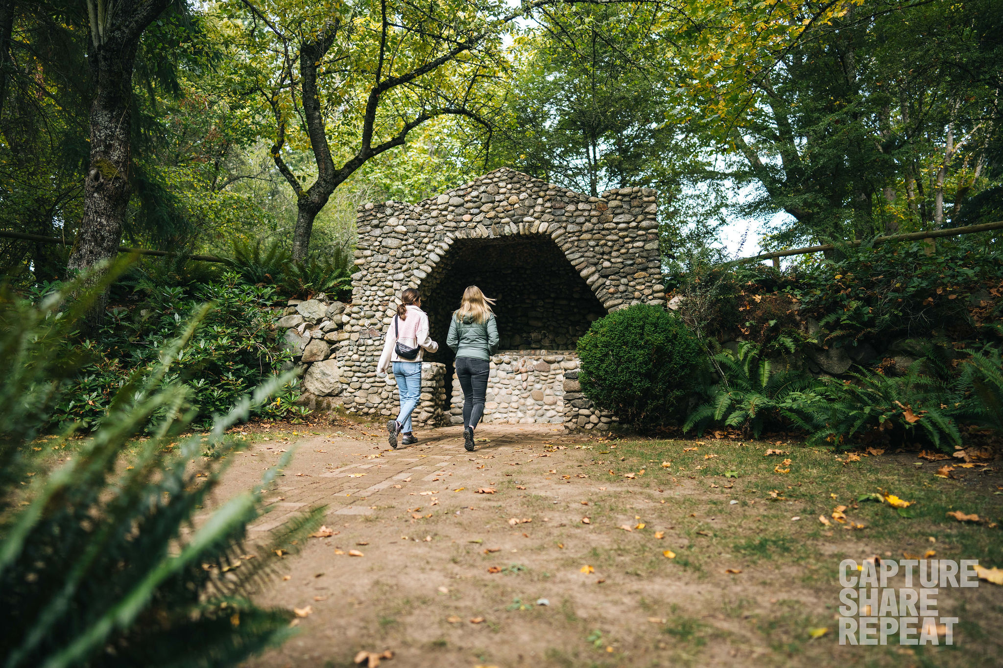 Two women walking up to a stone grotto with green trees surrounding the natural outdoor environment.