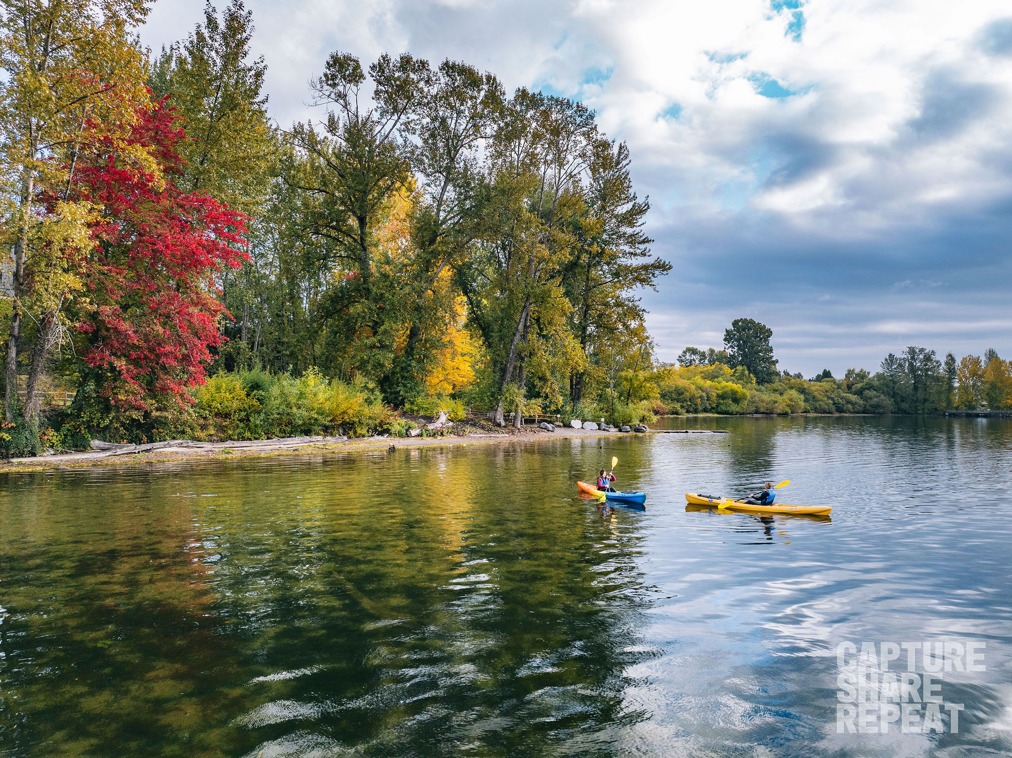 Two kayakers on a calm lake with fall foliage on the trees on the shoreline.