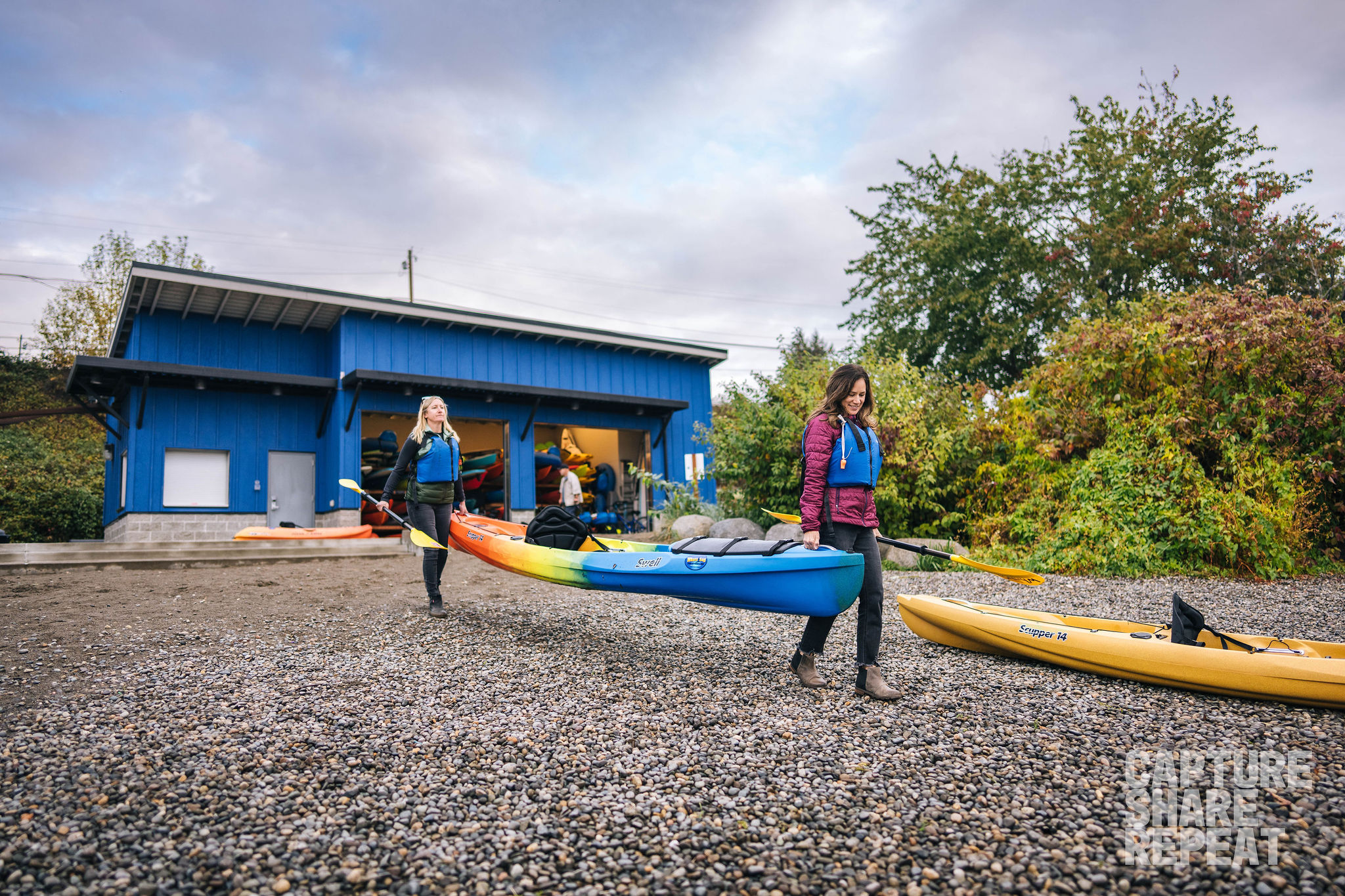 Two women carrying a kayak on the shoreline with a blue boathouse in the background.
