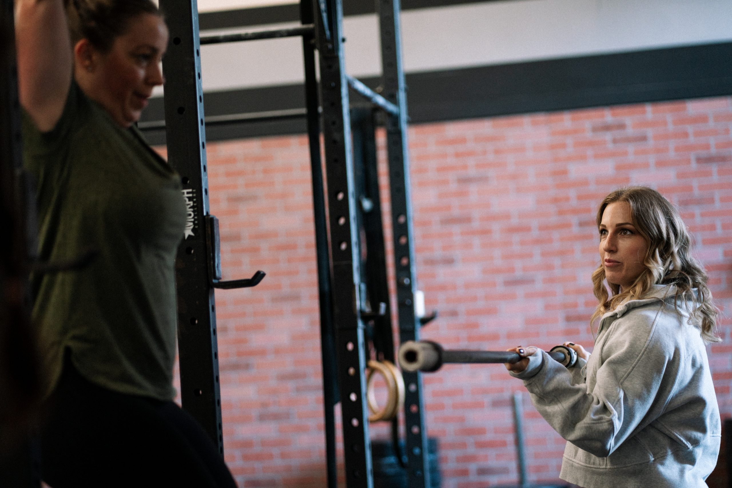 A woman in a gray sweatshirt holds a barbell while coaching another woman exercising in a gym with brick walls and weight racks.