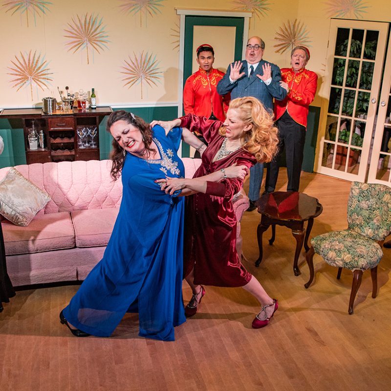 The set of a play in a living room with two ladies grabbing each other and others looking on surprised.